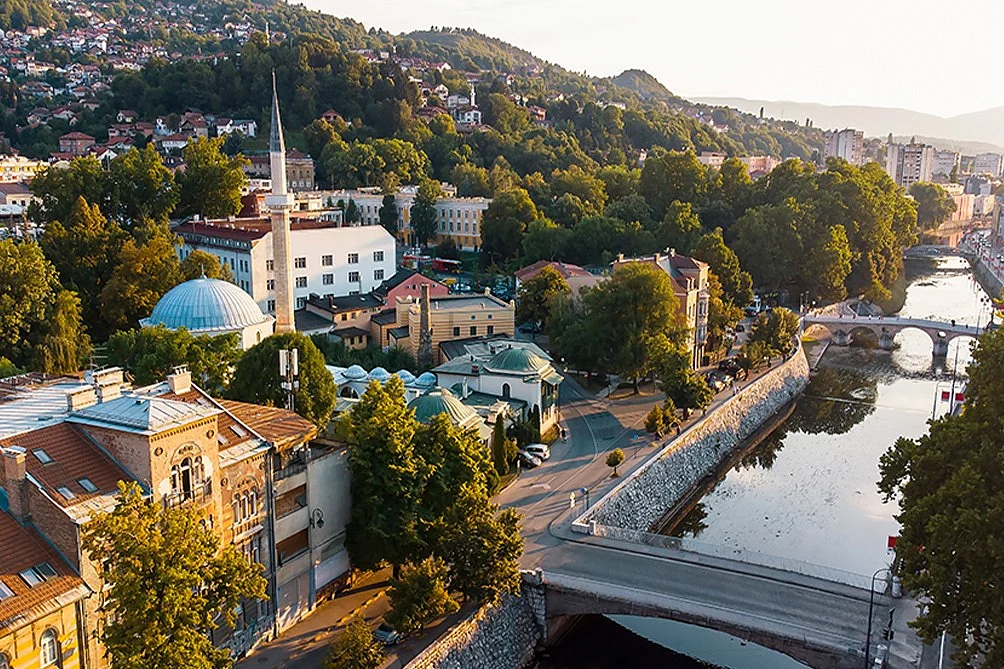A red cable car ascends over Sarajevo's dense cityscape toward lush green mountains under a bright blue sky.